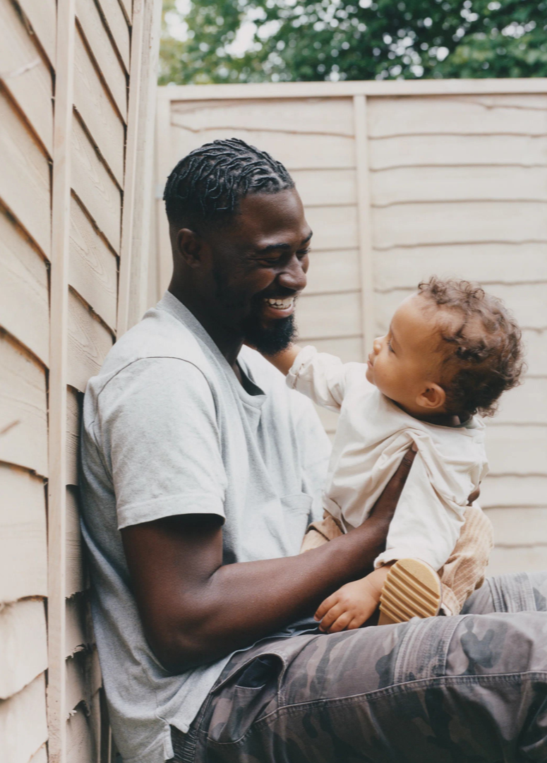 Man sitting outdoors with a child, leaning against a wooden wall.